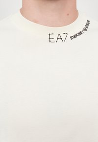Hvit bomulls t-skjorte med rund hals. Har svart brodert "EA7" og "Emporio Armani" merke ved halslinjen. Glatt tekstur.
