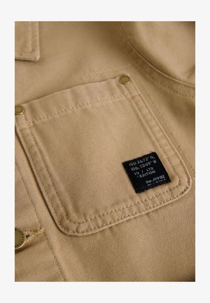 Béžová látka s texturovaným povrchem, která má náprsní kapsu s černou etiketu, mosazné patentky a kontrastní šicí detaily.