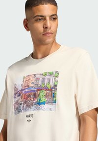 T-shirt od pamuka boje kreme s printanom ilustracijom scene pariškog kafića s plavim i zelenim naglascima, tekst "PARIS" ispod.