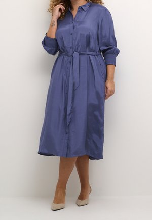 Vrouw draagt een midi-lang, blauw satijnen jurk met knopen, lange geplooide mouwen, een kraag, een tailleceintuur en beige puntige hakken.