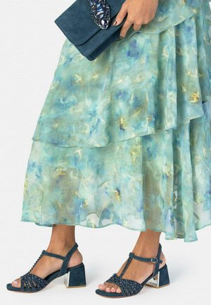 Mujer con falda en capas verde azulada, sosteniendo un bolso de mano azul oscuro con adornos y sandalias de tacón bloque azul oscuro con correas al tobillo.