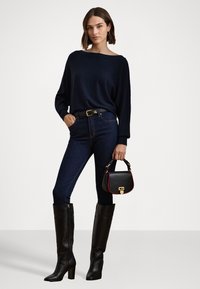 Fekete bőr térdcsizma, sötétkék pulóver és farmernadrág. Kis fekete kézitáska piros kiegészítőkkel. A modell magabiztosan áll.