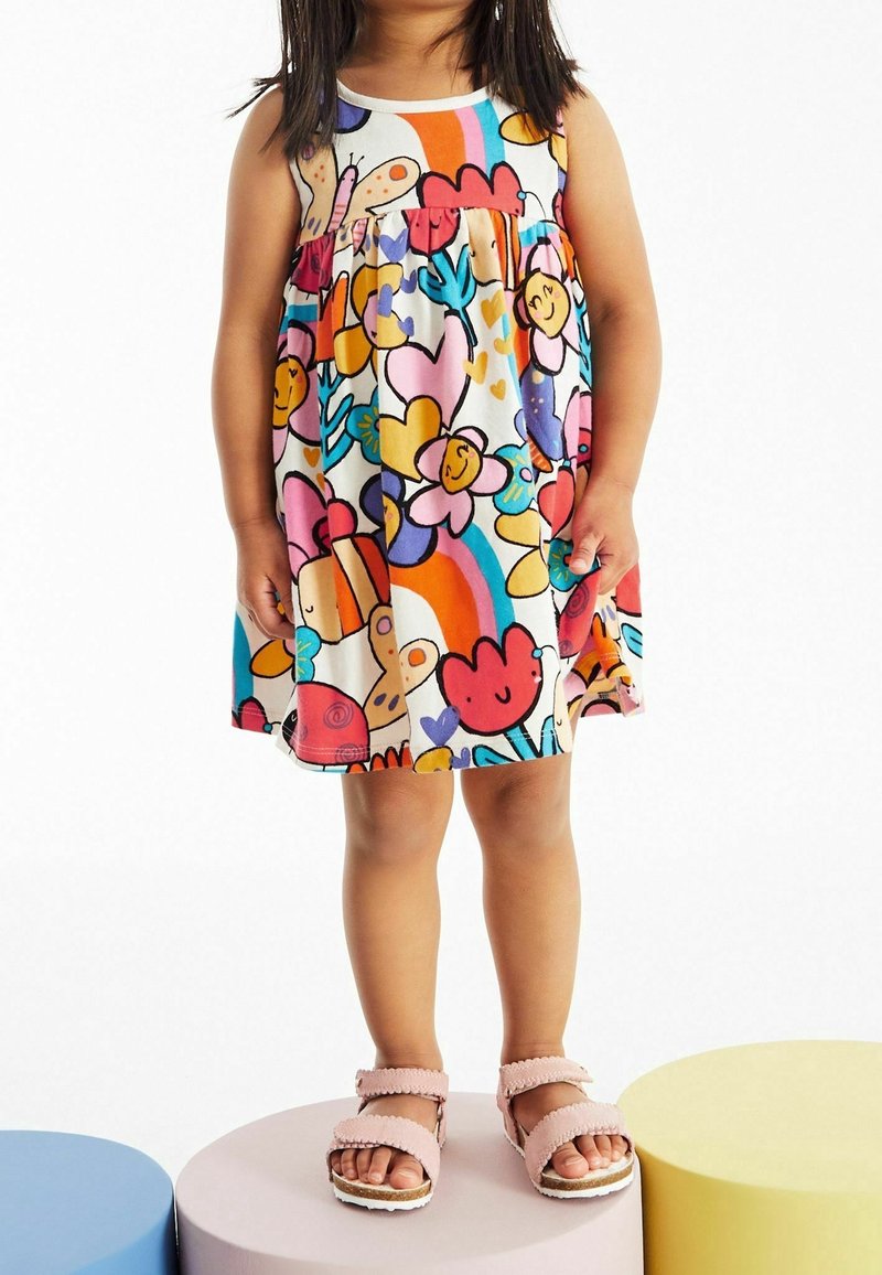 Enfant debout sur des plateformes rondes pastel portant une robe colorée avec des fleurs, des arcs-en-ciel et des papillons, ainsi que des sandales roses.