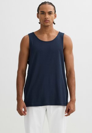 Joven con el pelo trenzado, vestido con una camiseta sin mangas azul marino y pantalones blancos, de pie frente a un fondo liso.