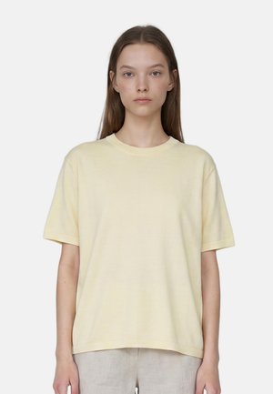 Jeune avec de longs cheveux portant un t-shirt jaune clair uni à manches courtes et un pantalon de couleur neutre, regardant devant avec une expression neutre.