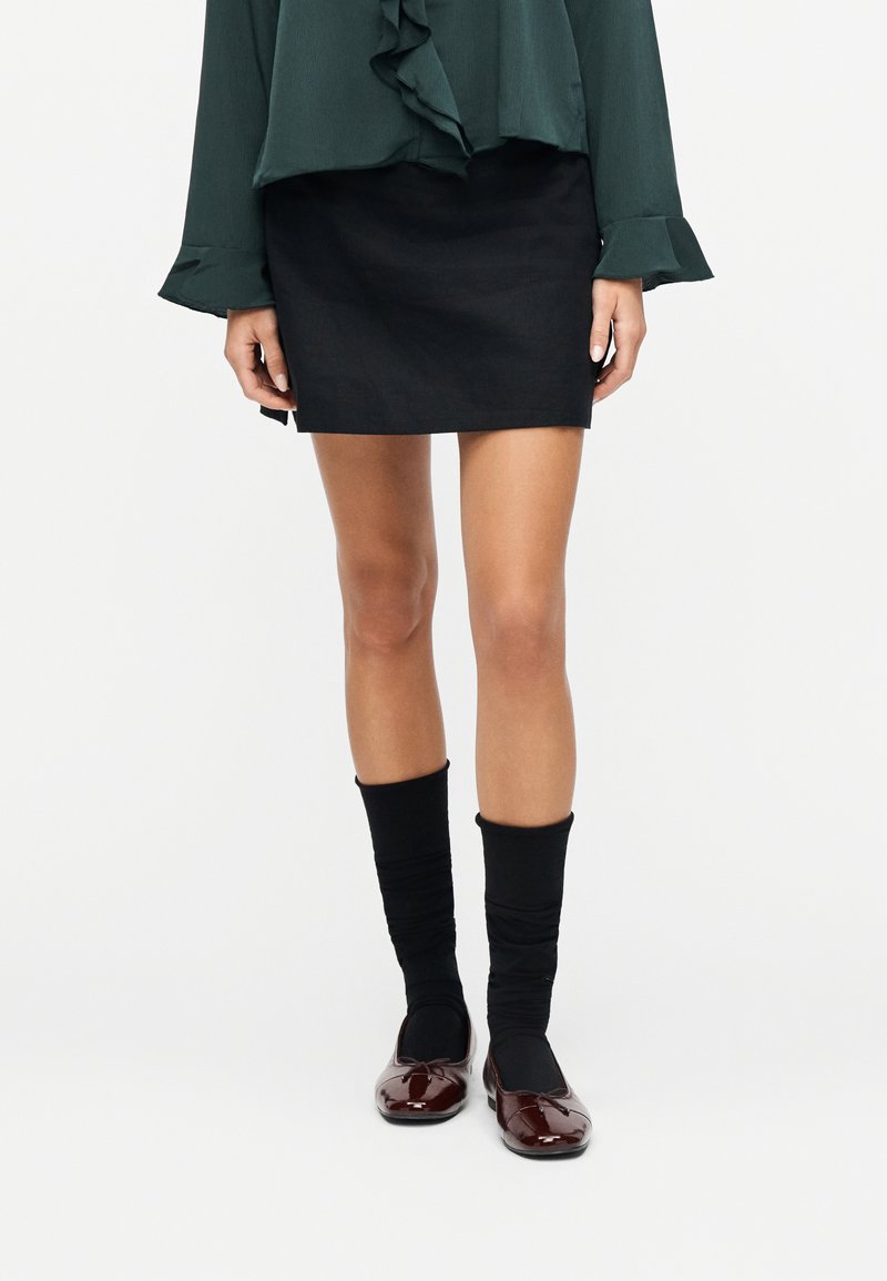 Femme portant un chemisier vert foncé à volants, une mini-jupe noire, des chaussettes noires mi-mollet et des ballerines brillantes bordeaux, debout devant un fond blanc.