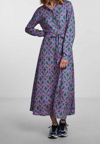 Lång knapp-up klänning med ett tyg i lila, rosa och gröna hjärtan, med knytmidja och långa ärmar, kombinerad med sportiga sneakers.