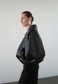 Chaqueta de cuero negra de gran tamaño con un gran cuello, bolsillos laterales y mangas estructuradas. El modelo lleva pendientes grandes y geométricos.