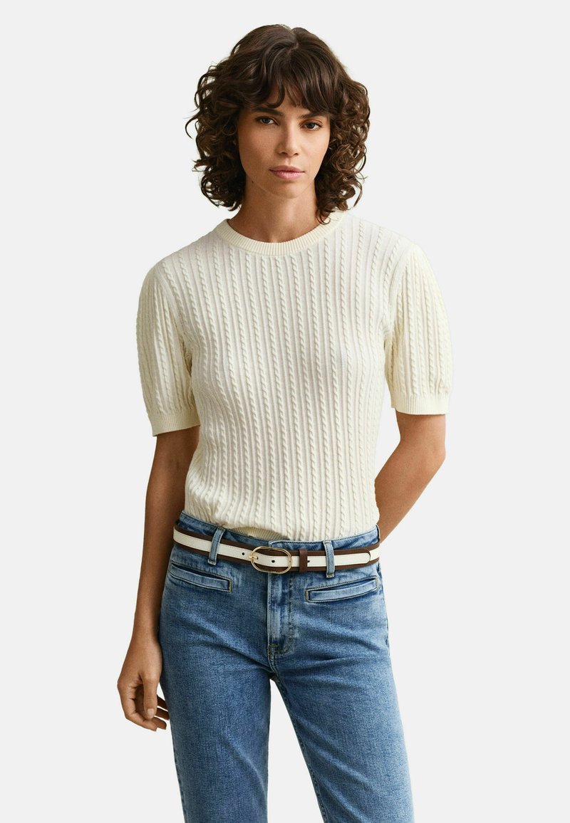 Mujer con cabello rizado que lleva un suéter blanco de punto cable de manga corta, jeans azules y un cinturón blanco con una franja marrón y hebilla dorada.