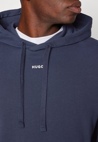 Homme portant un sweat à capuche bleu marine avec texte blanc "HUGO", regardant sur le côté, chemise blanche visible sous le sweat.