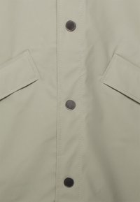 Light beige waterproof jacket with a smooth texture, featuring metallic snap buttons, flat pockets, and visible stitching along the front.