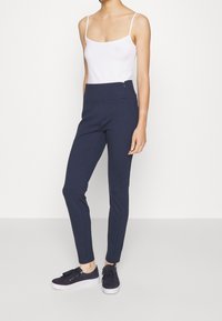 Pantalons slim-fit bleu marine foncé avec une taille élastique et un détail de fermeture éclair sur le côté, associés à un débardeur blanc et des baskets noires.