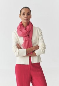 Leggero scialle rosa con orli sfrangiati, avvolto attorno a un cardigan color crema chiaro abbinato a pantaloni rosa, creando un look a strati.