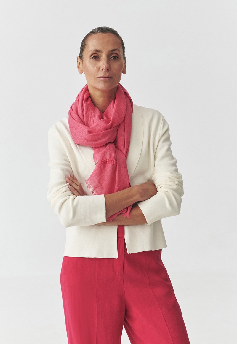 Leggero scialle rosa con orli sfrangiati, avvolto attorno a un cardigan color crema chiaro abbinato a pantaloni rosa, creando un look a strati.