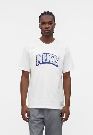 Ung mand med flettet hår iført hvid Nike t-shirt med blåt logo og grå bukser, stående mod en enkel hvid baggrund.