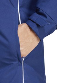 Marineblaue Jacke mit einer Reißverschlusstasche, die eine glatte Textur aufweist und kontrastierende weiße Einfassungen entlang der Nähte und Bündchen hat.
