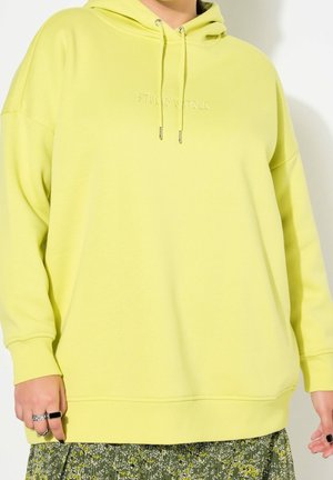 Personne portant un sweat à capuche oversize vert clair avec le texte "STUDIO UNTOLD" et une jupe à motif floral, montrant des mains avec des bagues et du vernis à ongles noir.