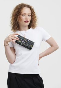 Black floral wallet with colorful wildflower pattern, held by a person wearing a white t-shirt and black pants. Smooth texture, rectangular shape.