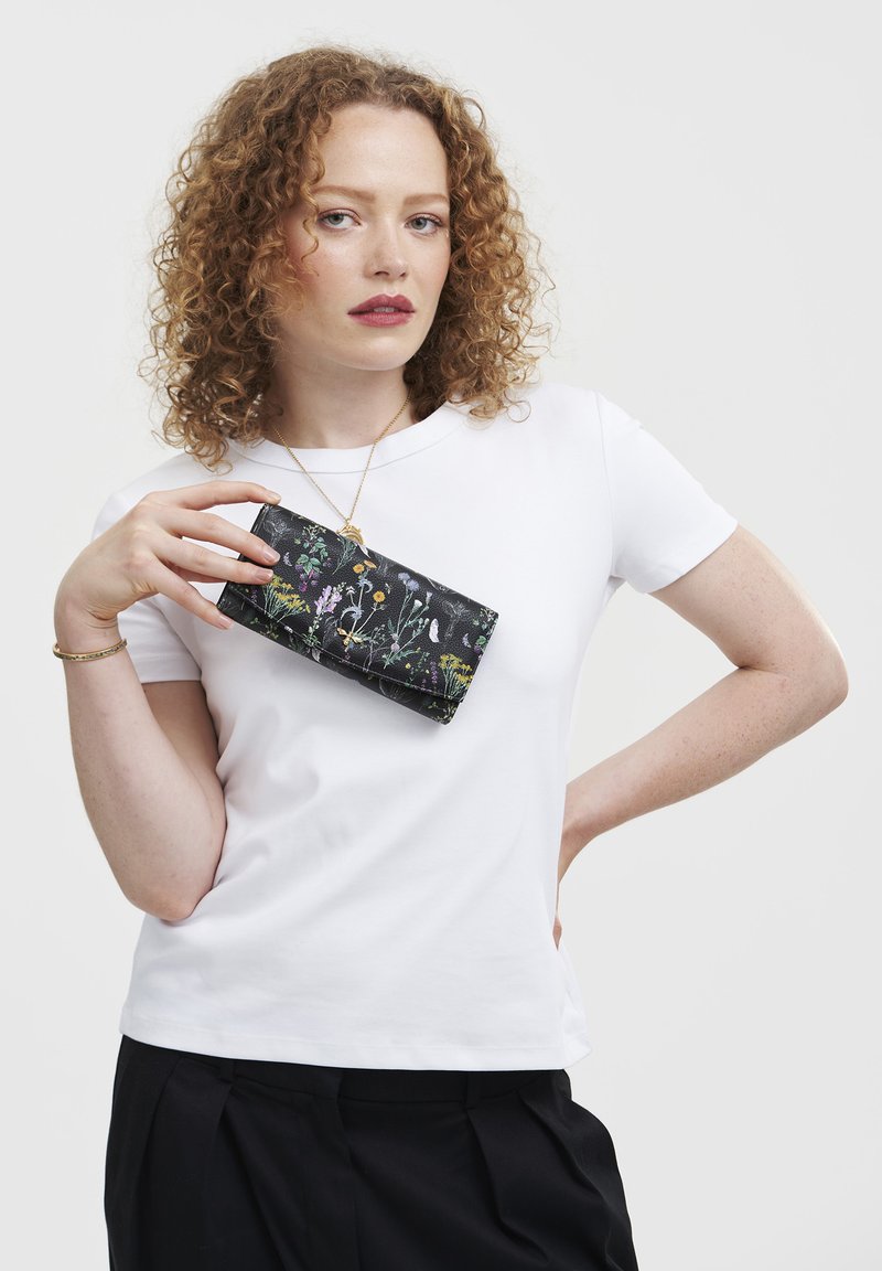 Black floral wallet with colorful wildflower pattern, held by a person wearing a white t-shirt and black pants. Smooth texture, rectangular shape.
