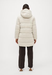 Polstret beige corduroy frakke med hætte, der har vandret søm og en længde til midt på låret. Sat sammen med mørkebrune corduroy bukser.