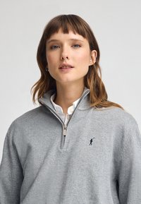 Pullover grigio con zip e colletto alto, realizzato in un morbido tessuto, caratterizzato da un piccolo logo scuro sul petto e una classica camicia bianca sotto.