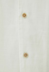 Camisa blanca de botones con un sutil patrón texturizado, que presenta un detalle de botón de madera en la parte superior, resaltando una tela ligera y transpirable.