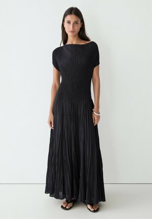 Donna con lunghi capelli scuri che indossa un abito maxi nero a maniche corte con texture e sandali neri, in piedi davanti a uno sfondo bianco uniforme.