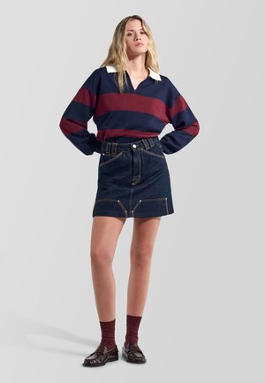 Donna che indossa una maglietta a maniche lunghe rigata blu navy e bordeaux, minigonna in denim scuro, calzini bordeaux e mocassini scuri, in piedi con le mani sui fianchi.