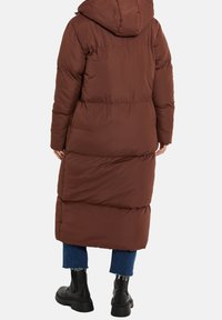 Abrigo largo acolchado marrón con capucha, con un diseño acolchado, puños elásticos y un corte relajado, combinado con jeans de mezclilla azul y botas negras.