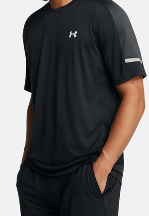 Homme portant un t-shirt de sport noir Under Armour à manches courtes avec logo sur la poitrine et main dans la poche sur fond uni.