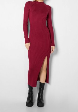 Abito bordeaux a costine con maniche lunghe e spacco laterale, caratterizzato da una silhouette aderente e un collo alto. Abbinato a stivali neri con tacco alto.
