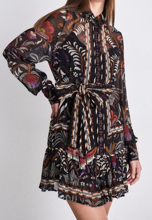 Frau trägt ein langärmliges, schwarzes Kleid mit floralen Mustern in Orange, Weiß und Lila, mit Knopfleiste vorne und einem gebundenen Taillengürtel.