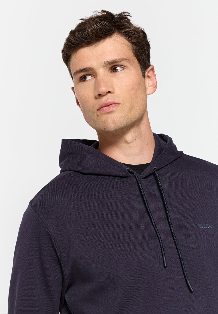 Le hoodie bleu marine présente un tissu texturé, des poignets côtelés, un cordon de serrage réglable et un logo discret sur la poitrine.