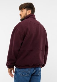 Burgundy színű bolyhos dzseki magas gallérral, texturált felülettel, rugalmas aljjal és oldalsó varrási részletekkel. A modell hátulról látható.