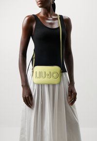 Sac bandoulière rectangulaire vert clair avec logo "LIU JO" embossé. Texture en cuir lisse, bandoulière ajustable, associé à un débardeur noir et une jupe blanche.