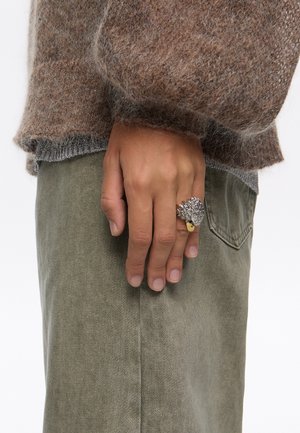 Anillo de plata con un diseño texturizado que recuerda a una hoja, llevado en una mano junto a pantalones de color verde oliva. El suéter es de un suave tejido marrón con un corte holgado.