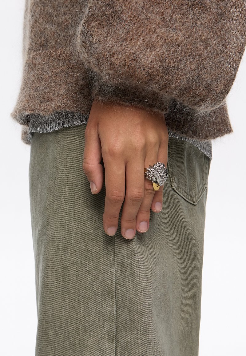 Anillo de plata con un diseño texturizado que recuerda a una hoja, llevado en una mano junto a pantalones de color verde oliva. El suéter es de un suave tejido marrón con un corte holgado.