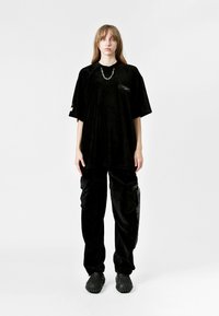 Camiseta oversized de corduroy negra con mangas cortas y un pequeño bolsillo en el pecho, combinada con pantalones cargo negros holgados que tienen bolsillos laterales.