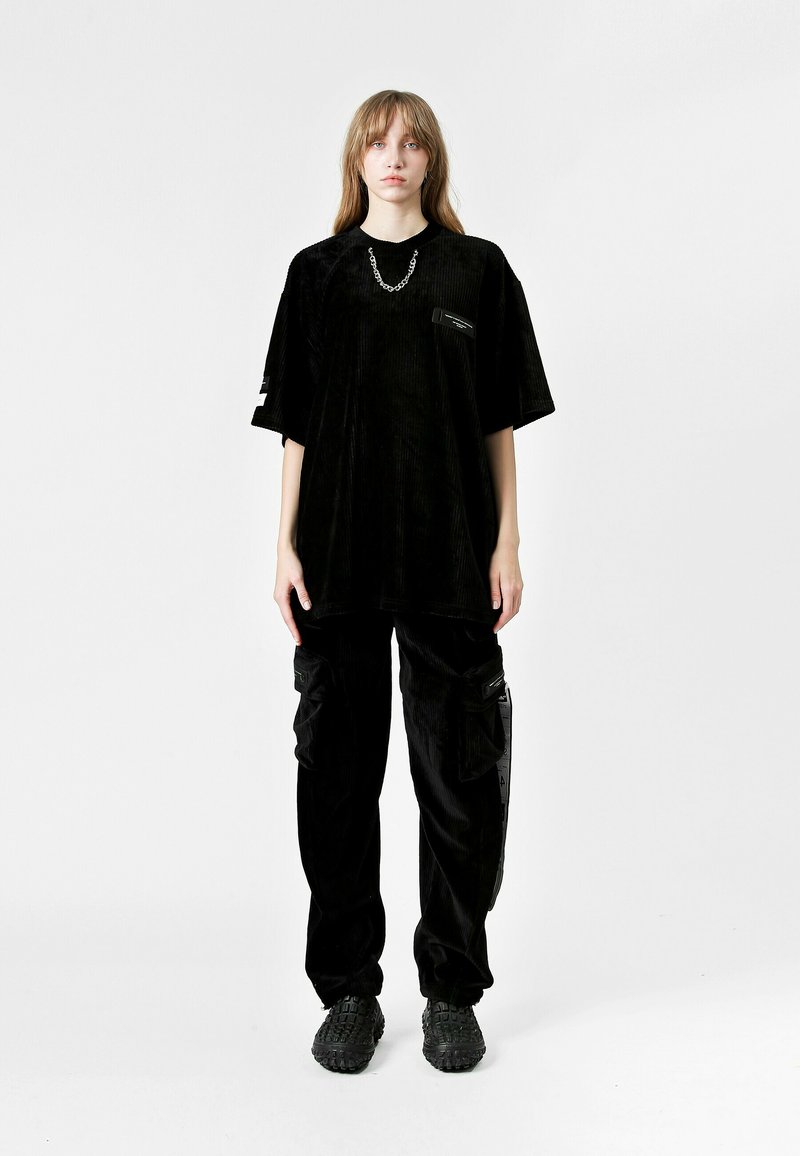 Camiseta oversized de corduroy negra con mangas cortas y un pequeño bolsillo en el pecho, combinada con pantalones cargo negros holgados que tienen bolsillos laterales.