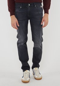 Uomo che indossa jeans scuri, sneakers bianche e beige con dettagli blu, e un maglione borgogna, in piedi contro uno sfondo chiaro e semplice.