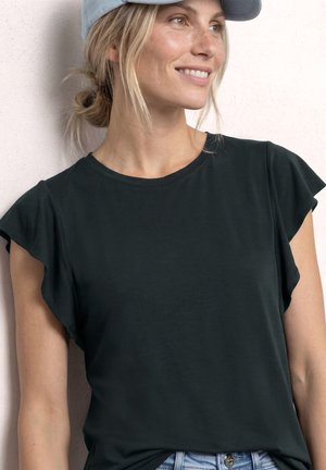 Smilende kvinde med blondt hår i en lav knold iført en lyseblå kasket, sort bluse med flæser på ærmerne og lyseblå jeans, kigger til siden.