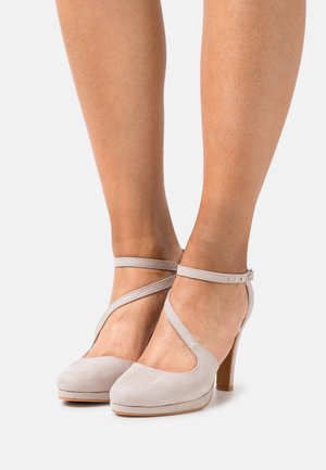 Jambes d'une femme portant des escarpins en daim gris clair avec des straps de cheville croisés et une fermeture à boucle sur un fond blanc.