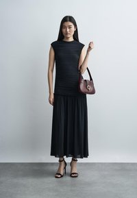 Vrouw in een mouwloze zwarte jurk, zwarte sandalen met hoge hakken, met een donkerrode handtas, staande tegen een effen lichtgrijze achtergrond.