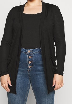Kvinde iført sort cardigan med åben front over sort top og høj taljede blå jeans med fire synlige knapper mod ensfarvet baggrund.
