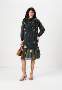 Robe noire transparente avec un col à volants, des manches longues et un ourlet en couches, ornée d'imprimés animaliers et de feuilles multicolores. Portée avec des talons à lanières.
