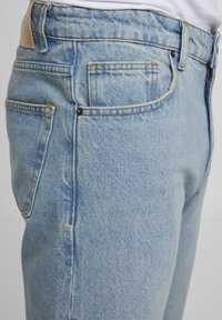 Világoskék farmernadrág látható első zsebbel, övbújtatókkal és narancssárga varrással, semleges háttér előtt.