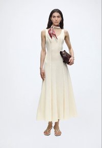 Γυναίκα σε μπεζ midi πλεκτό φόρεμα χωρίς μανίκια, κόκκινο ριγωτό φουλάρι στον λαιμό, μπεζ σαγιονάρες, που κρατά καφέ σκούρο τσαντάκι.