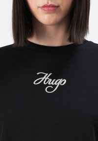 Close-up van een persoon met een zwarte top waarin "Hugo" in witte cursieve letters op de borst is geborduurd.