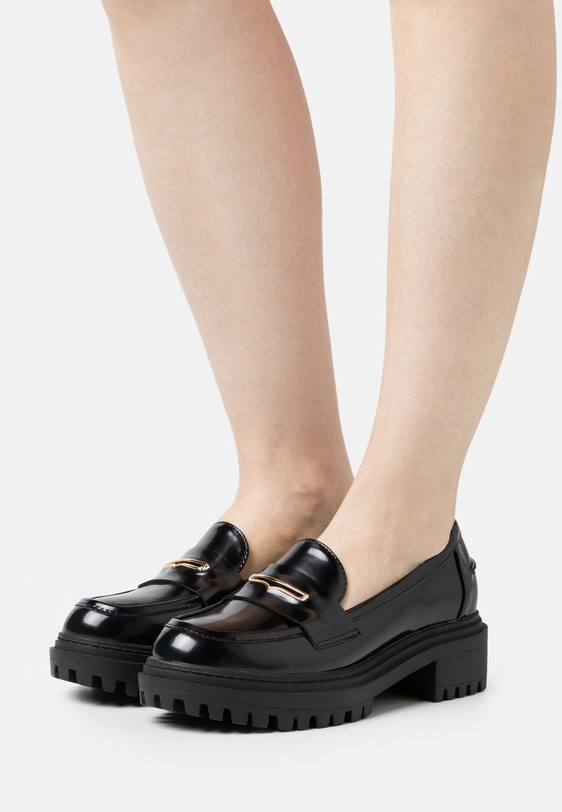 Mocassins en cuir noir brillant avec semelles compensées à crampons épais et accents métalliques dorés portés pieds nus sur un fond blanc.