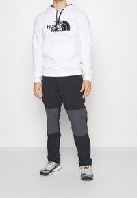 Vit hoodie med svart logotyp, i kombination med svarta byxor som har grå detaljer. Modellen bär grå och svarta sneakers. Enkel och funktionell design.